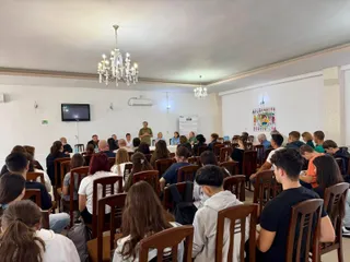 Un grup de persoane este așezat pe scaune de lemn în fața unui bărbat care stă în picioare într-o încăpere cu candelabre și un televizor.