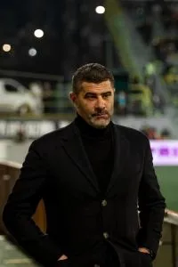 Un bărbat cu părul scurt și barbă căruntă poartă un sacou negru peste un tricou cu guler înalt negru, stând cu mâinile în buzunare.