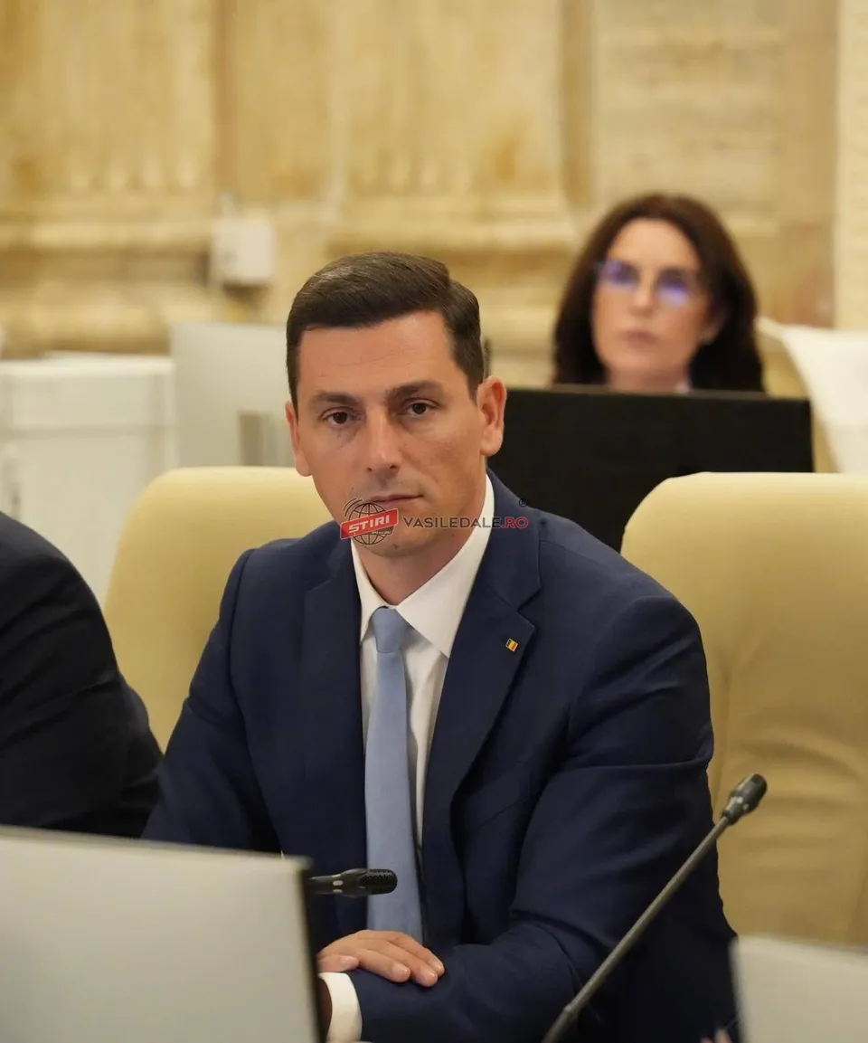 Un bărbat în costum, cu cravată, stă la o masă, privind înainte, cu o femeie în fundal.