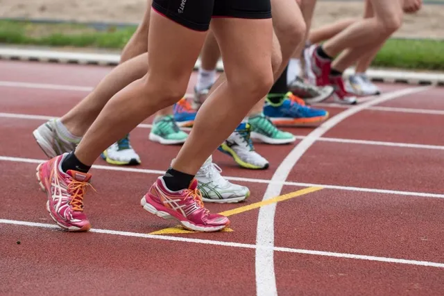 Picioarele unor atleți în echipament sportiv și încălțăminte colorată sunt poziționate pe o pistă de atletism marcată cu linii albe și galbene.