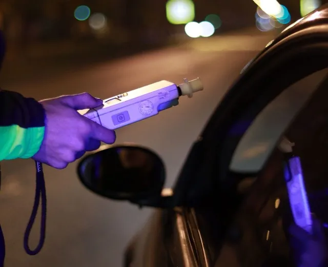 O mână ține un aparat alb de testare alcoolemiei în apropierea unei mașini în timpul nopții.