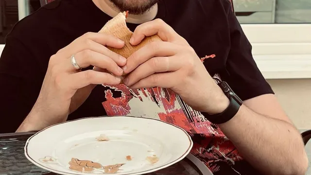 Un bărbat cu o cămașă neagră și un ceas de mână ține un sandviș cu ambele mâini deasupra unei farfurii albe cu resturi de mâncare.