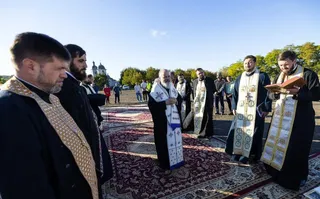 Câțiva bărbați îmbrăcați în haine negre și cu stole albe ornate stau în picioare pe un covor decorativ colorat sub cerul senin.