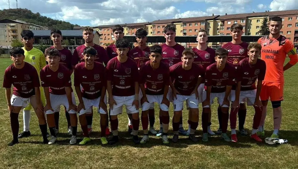 Un grup de fotbaliști tineri în echipamente de sport de culoare bordo și portocalie pozează într-o linie pe un teren de fotbal cu iarbă.