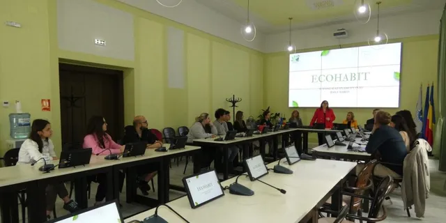 Un grup de persoane stă la mese lungi într-o sală cu pereți verzi, privind un ecran care afișează textul ECOHABIT.