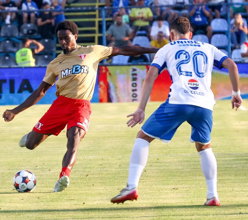 Un jucător de fotbal în echipament galben și roșu lovește mingea pe un teren de iarbă în fața unui adversar în echipament alb și albastru.