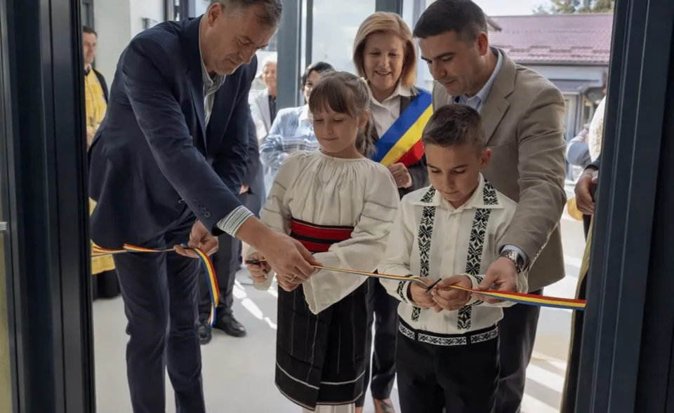 Un grup de adulți și doi copii în haine tradiționale taie o panglică colorată la o intrare cu uși de sticlă.