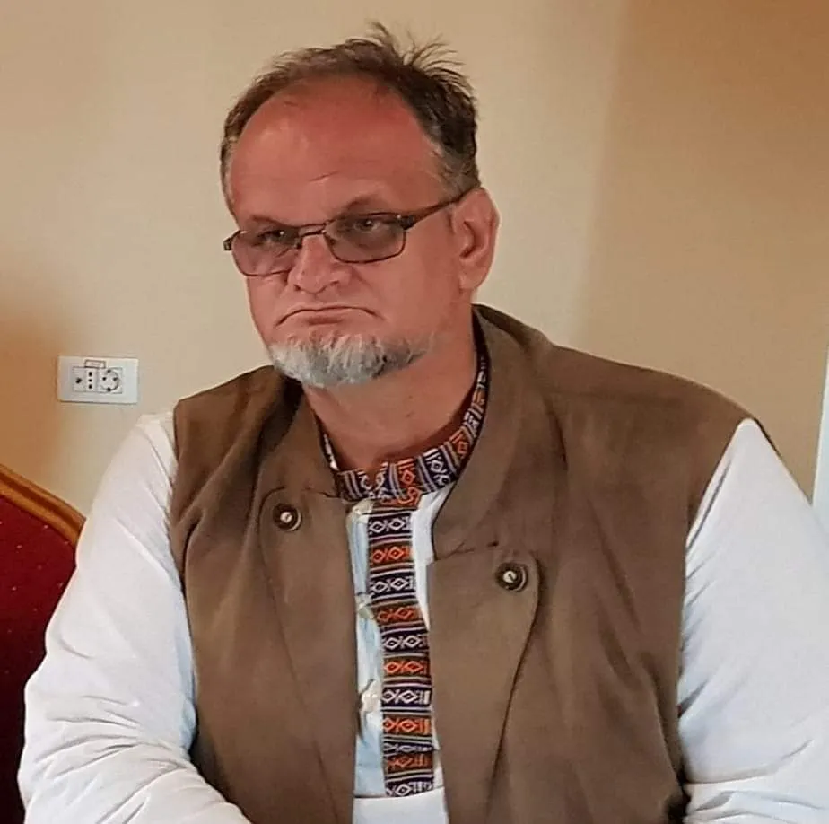 Un bărbat cu ochelari, purtând o vestă maro peste o cămașă albă cu un guler decorat, privește spre lateral.