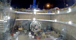 Două utilaje de forare circulare sunt poziționate pe fundul unui puț de construcție de beton, sub iluminare artificială nocturnă.