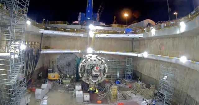 Două utilaje de forare circulare sunt poziționate pe fundul unui puț de construcție de beton, sub iluminare artificială nocturnă.