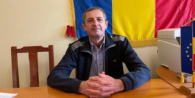 Un bărbat cu o cămașă în dungi și un hanorac închis la culoare stă la o masă de lemn în fața unui steag colorat.