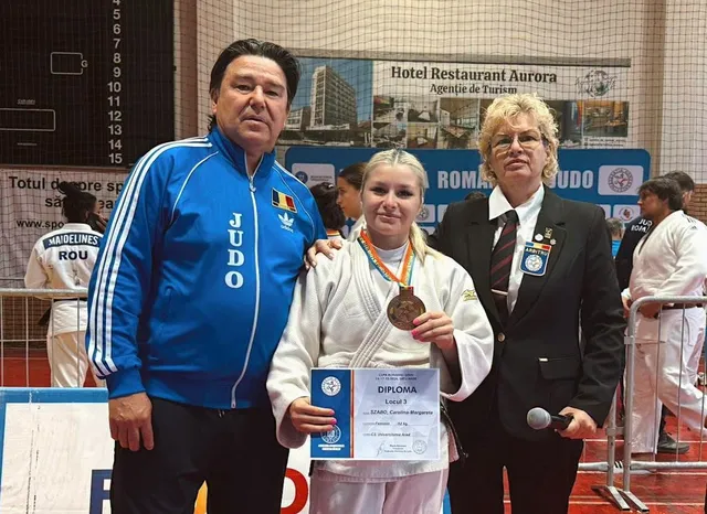 Un bărbat în costum sport albastru, o femeie în halat alb cu medalie și un certificat, și o femeie în costum negru stau împreună.