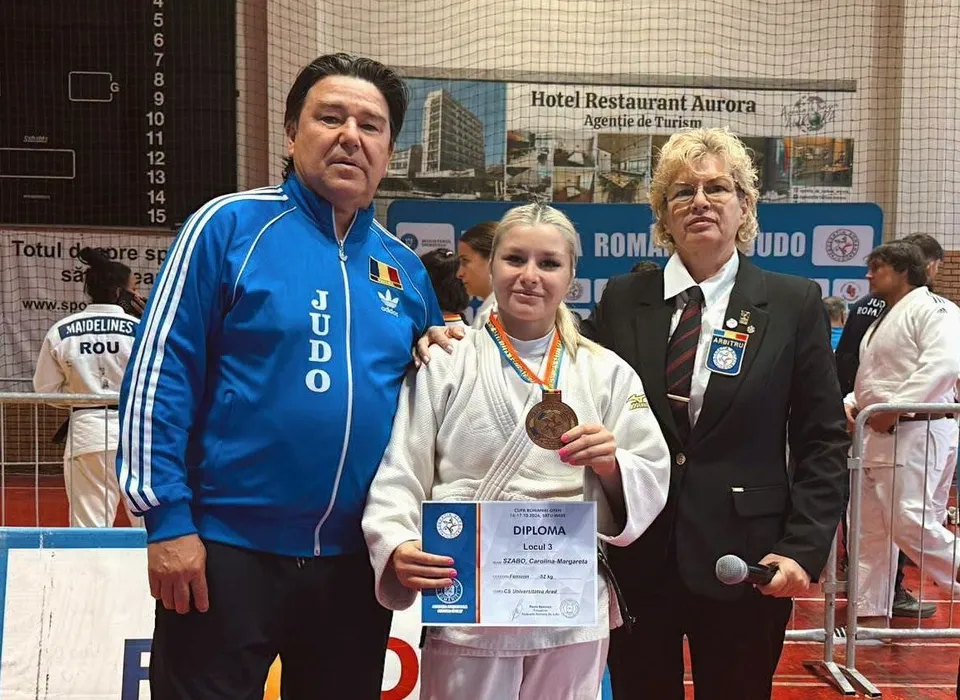 Un bărbat în costum sport albastru, o femeie în halat alb cu medalie și un certificat, și o femeie în costum negru stau împreună.