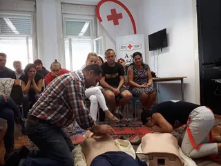 Un bărbat într-un cămașă în carouri este genunchiat lângă un manechin de simulare a resuscitării, în timp ce alți oameni stau așezați în cameră.