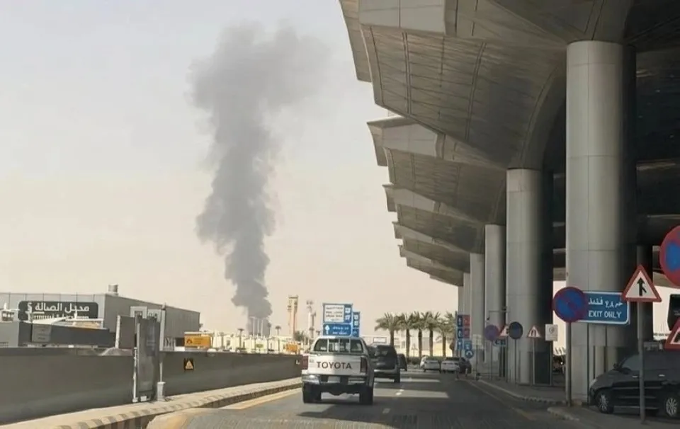 O coloană de fum negru se ridică deasupra unor clădiri într-un peisaj urban, lângă o structură de terminal cu coloane gri și un vehicul alb.