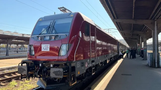 O locomotivă roșie cu logoul ARF stă pe un binar lângă un peron de gară sub un cer senin.