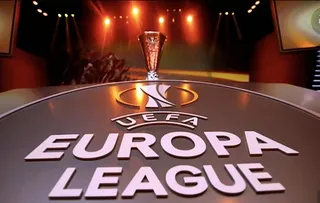 Un trofeu metalic este poziționat pe o suprafață plană în fața unui logo cu textul UEFA Europa League sub lumini de scenă.