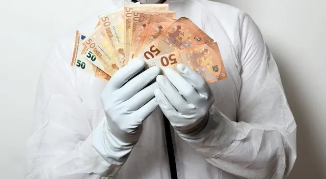 O persoană îmbrăcată într-un halat alb și mănuși medicale de protecție ține un teanc de bancnote de cincizeci de euro.