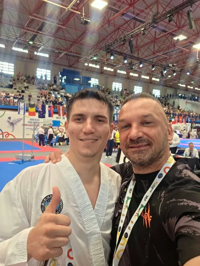 Un bărbat într-un costum de karate alb cu un deget ridicat și un bărbat într-un tricou negru zâmbesc în prim-plan într-o sală de sport.