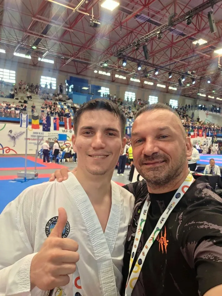 Un bărbat într-un costum de karate alb cu un deget ridicat și un bărbat într-un tricou negru zâmbesc în prim-plan într-o sală de sport.