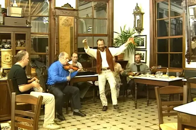 Un bărbat cu vestă maro și cămașă albă dansează cu brațele ridicate într-o încăpere cu ferestre mari, în timp ce alți bărbați ascultă muzică.