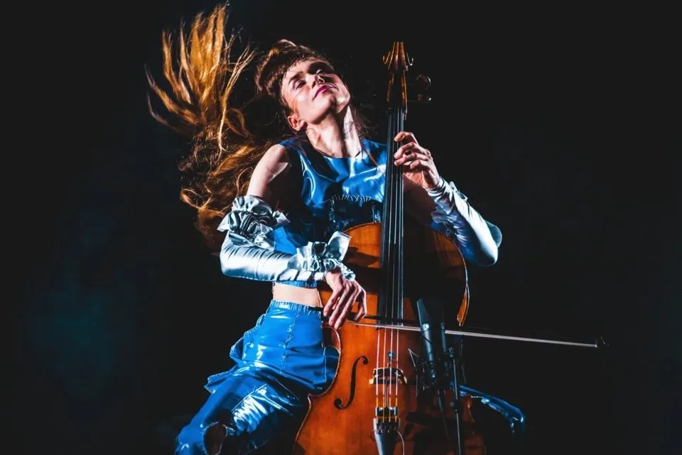 O femeie cu părul lung și mișcat de vânt cântă la un instrument de muzică cu coarde pe un fundal întunecat.