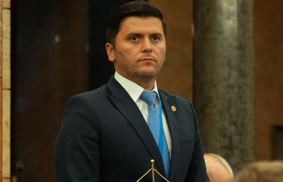 Un bărbat cu părul scurt, purtând un costum închis la culoare, cămașă albă și cravată albastră, stă în fața unui fundal de coloană de piatră.