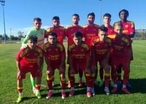 O echipă de fotbaliști în echipamente roșii și galbenii stă poziționată pe un teren de fotbal sub un cer senin.