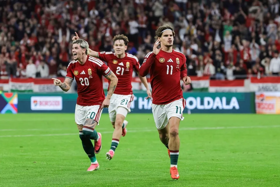 Trei jucători de fotbal în echipamente roșii și albi aleargă pe un teren de fotbal verde în fața unui public numeros.