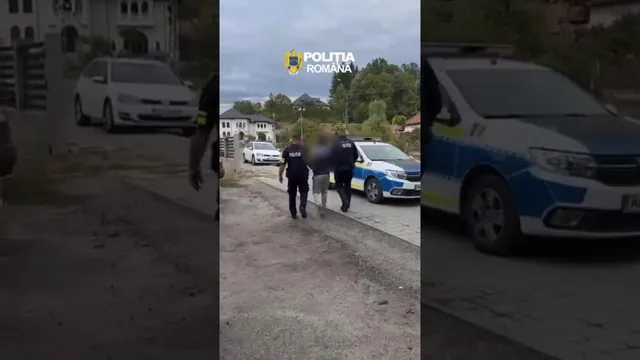 Trei persoane în uniformă întunecată stau lângă două mașini de poliție parcate pe un drum de pietriș lângă o clădire.