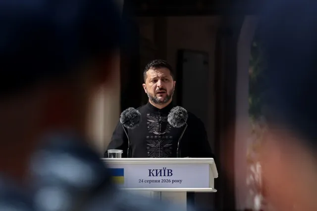 Un bărbat cu barbă și haină neagră stă în spatele unui pupitru alb pe care scrie text în alfabet chirilic.
