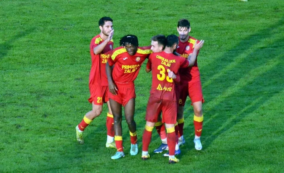 Cinci jucători de fotbal îmbrăcați în echipamente roșii și galbene stau grupați pe un teren de gazon verde.