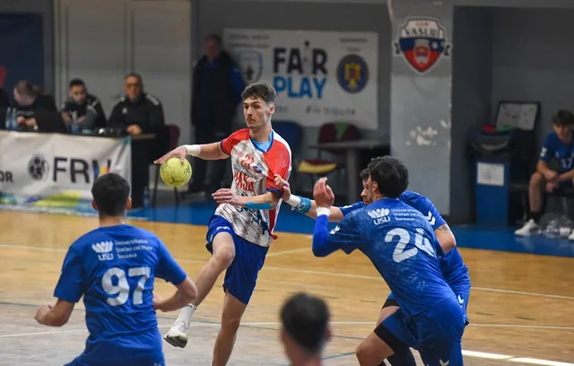 Un jucător de handbal în echipament alb și roșu ține o minge galbenă în timp ce este marcat de adversari în echipament albastru.