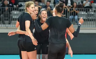 Cinci femei cu haine negre se îmbrățișează pe un teren de sport, în fața unor spectatori așezați pe scaune în tribune.