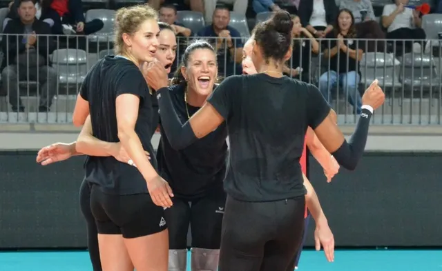 Cinci femei cu haine negre se îmbrățișează pe un teren de sport, în fața unor spectatori așezați pe scaune în tribune.