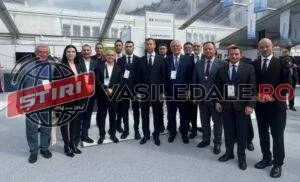Un grup de bărbați și o femeie în haine formale stau în rând pe o suprafață asfaltată în fața unei clădiri cu acoperiș metalic.