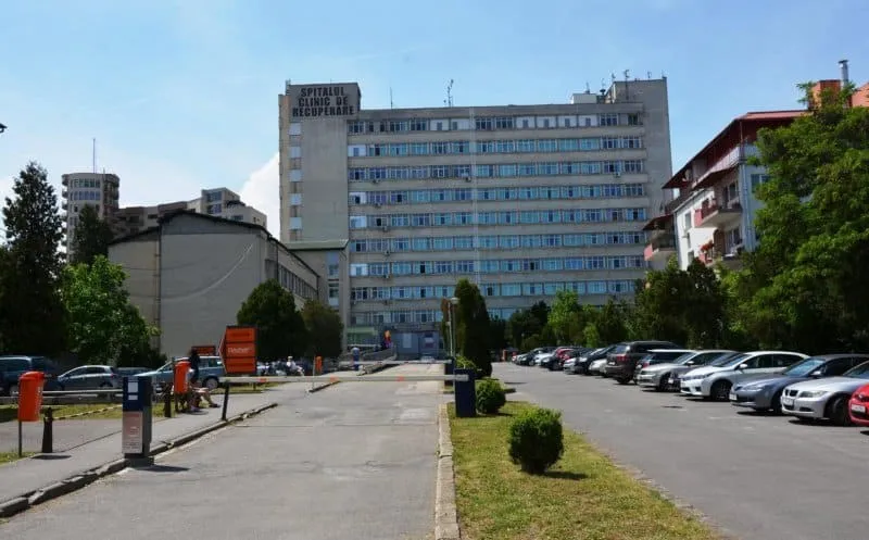 O clădire înaltă de beton cu multe ferestre este situată lângă un drum asfaltat pe marginea căruia sunt parcate mai multe mașini albe și gri.