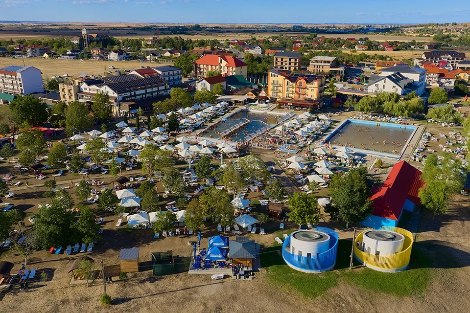 O vedere aeriană a unei zone cu piscine, umbrele și diverse clădiri de cazare înconjurate de vegetație și câmpuri deschise sub un cer senin.