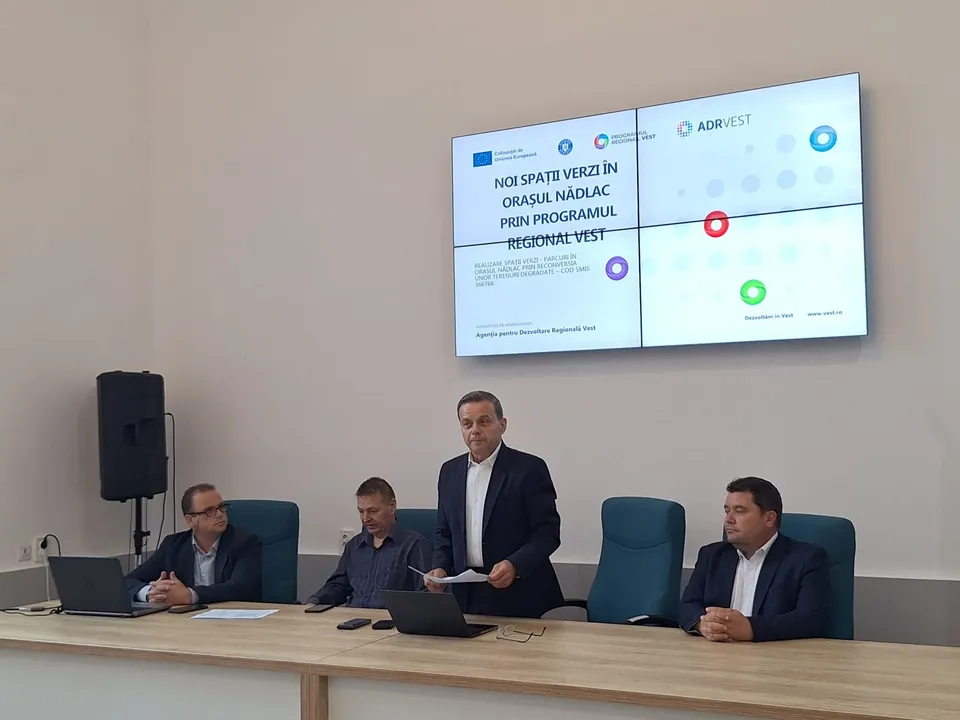 Patru bărbați stau la o masă lungă de lemn în fața unui ecran mare care afișează un text și câteva logo-uri.