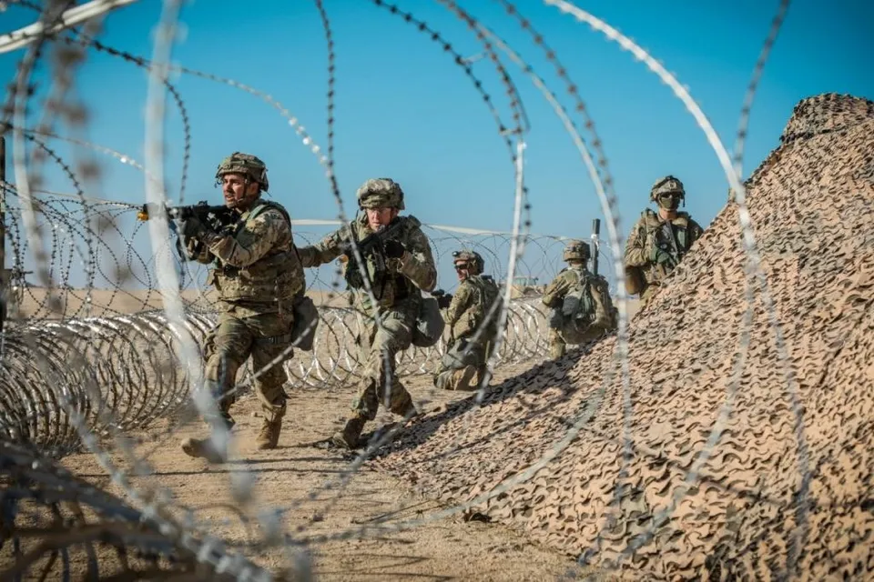 Cinci soldați în uniforme de camuflaj și echipament militar se deplasează printre garduri de sârmă ghimpată într-un teren deschis sub un cer senin.