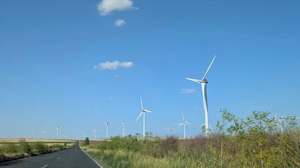 Mai multe turbine eoliene albe sunt amplasate pe un câmp verde sub un cer albastru cu câteva nori plutind.