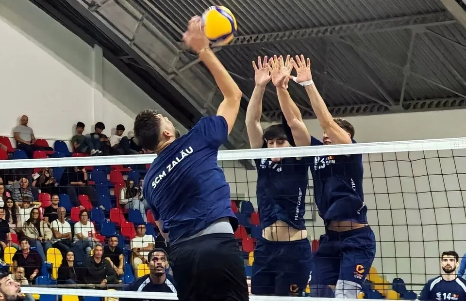 Un jucător de volei lovește mingea deasupra unei rețele în fața a doi adversari care ridică brațele pentru a bloca.