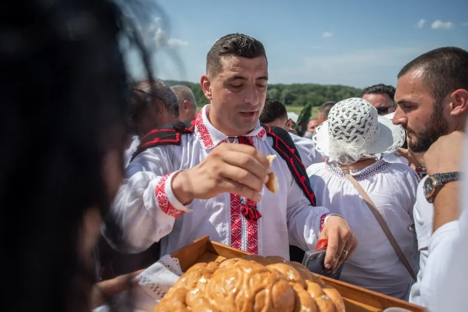 Un bărbat într-un cămașă tradițională albă cu broderii roșii ține o bucată de pâine deasupra unei tăvi cu cozonac într-un grup de oameni.