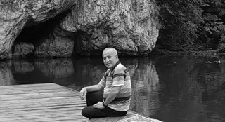 Un bărbat cu părul scurt și tricou polo stă așezat pe un ponton de lemn lângă un lac, având o stâncă și pădure în fundal.