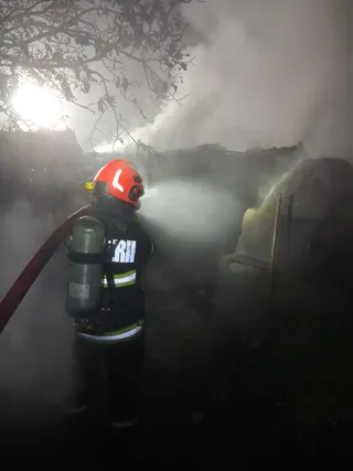 Un pompier echipat cu cască roșie, costum de protecție și oxigen purcește un jet de apă într-un mediu înfășurat în fum, probabil în timpul unui incendiu.
