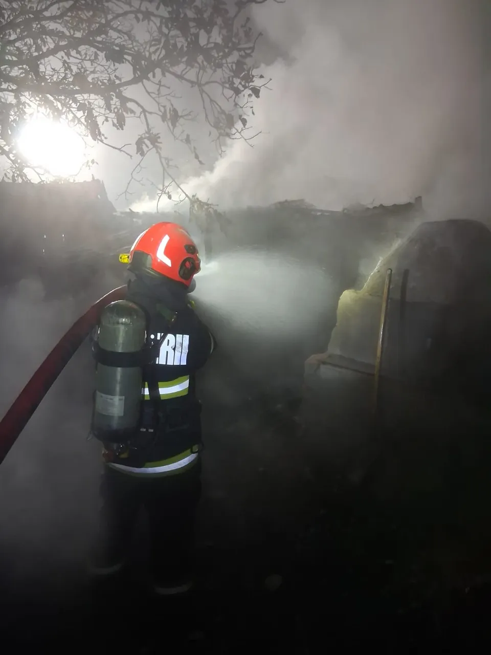 Un pompier echipat cu cască roșie, costum de protecție și oxigen purcește un jet de apă într-un mediu înfășurat în fum, probabil în timpul unui incendiu.
