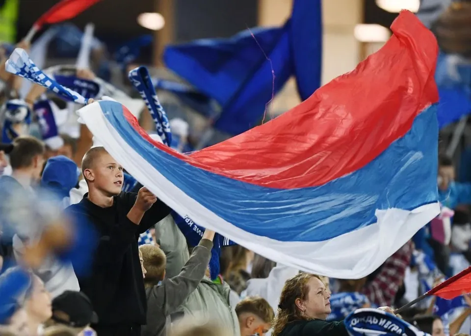 Un băiat cu părul scurt ține o steag tricolor cu benzi roșu, albastru și alb într-un grup de oameni.
