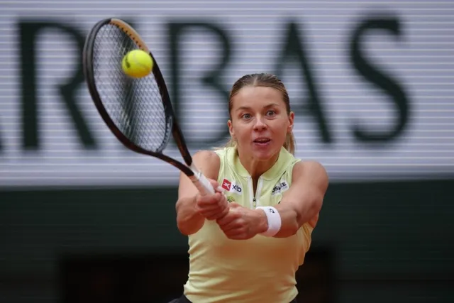 O femeie cu părul blond și tricou galben lovește o minge de tenis cu o rachetă pe un teren de tenis.
