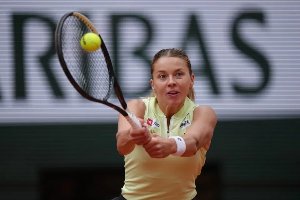 O femeie cu părul blond și tricou galben lovește o minge de tenis cu o rachetă pe un teren de tenis.