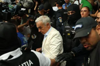 Un bărbat cu părul alb și sacou deschis este înconjurat de persoane în uniformă și de microfoane într-un spațiu aglomerat.